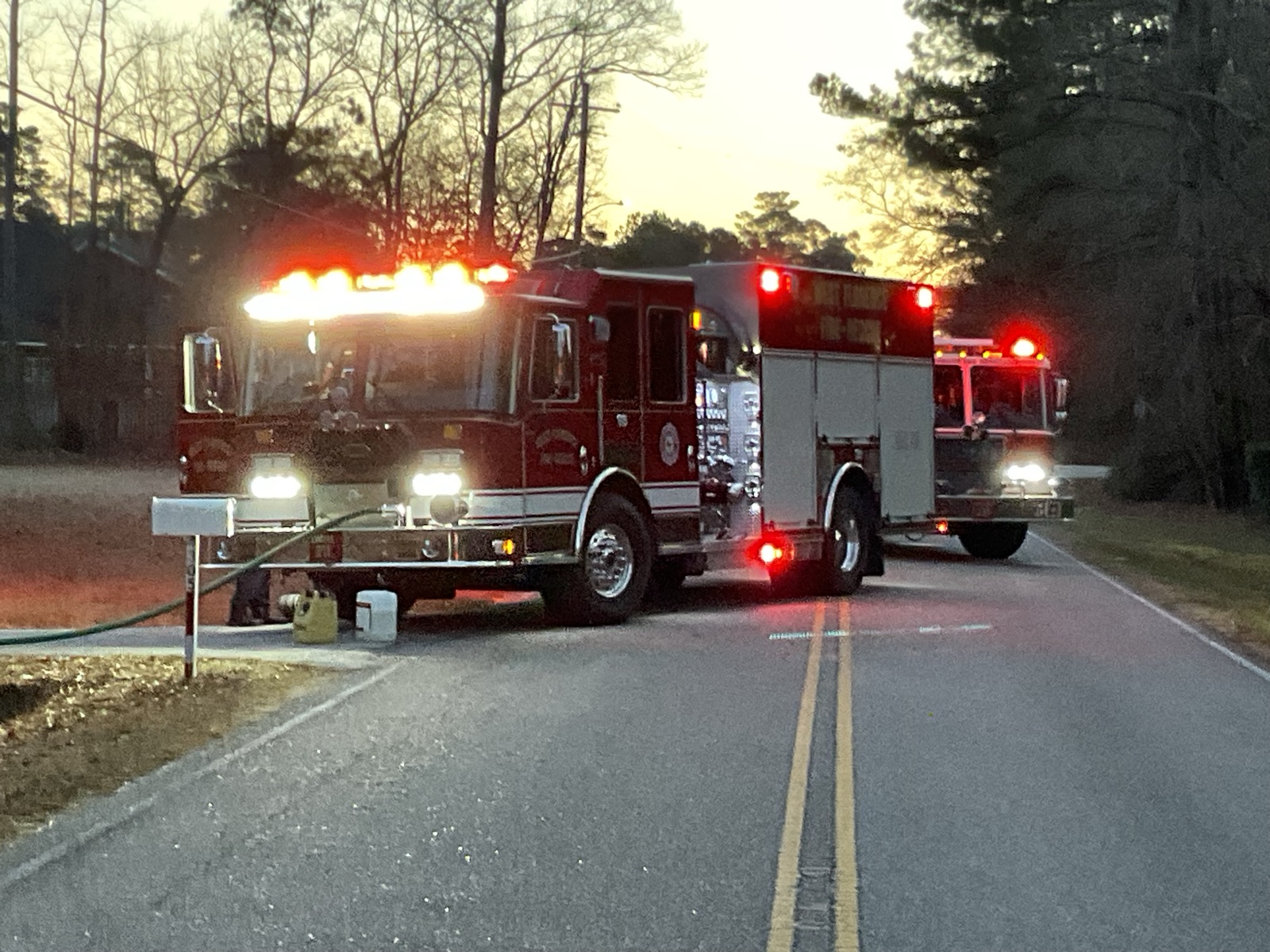 Two engines staged on a rural road at dusk with hose deployed and warning lights on